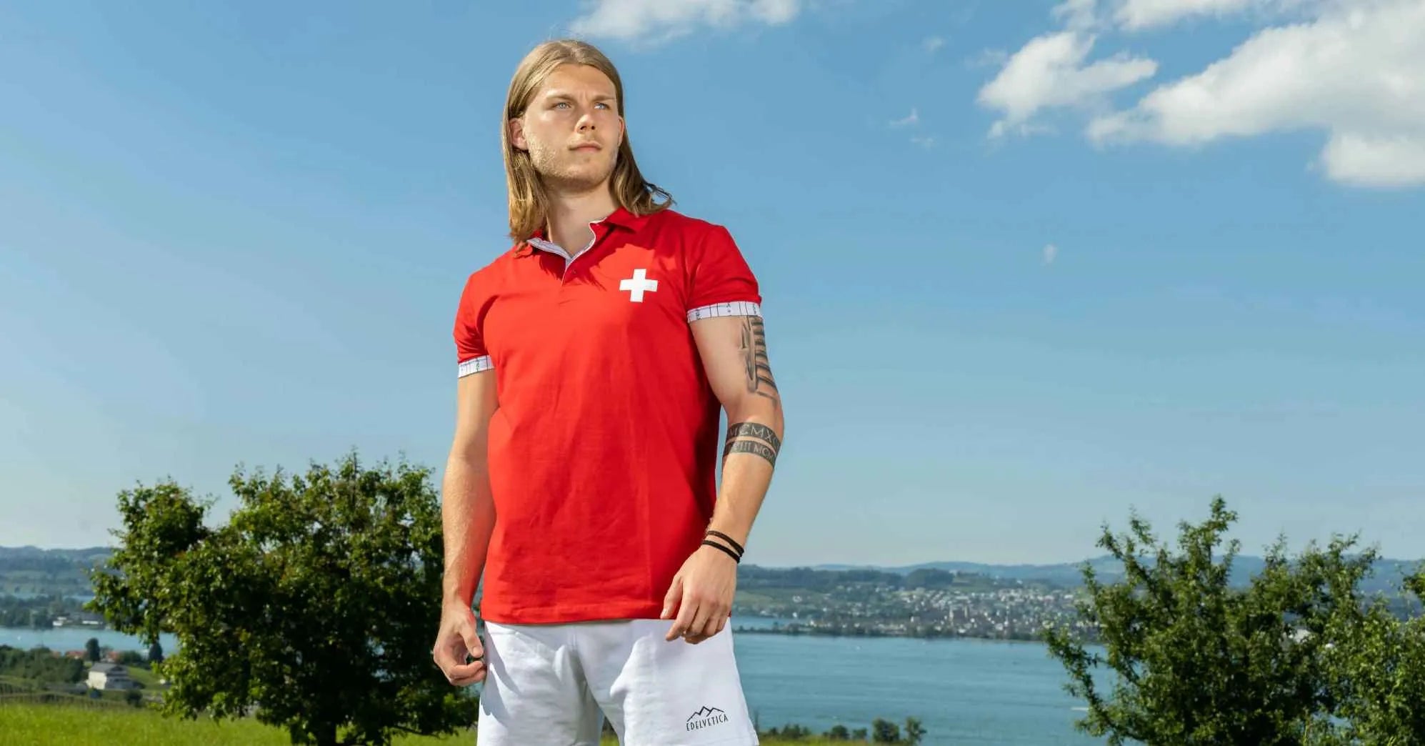 Junger Mann im roten Shirt mit Schweizer Kreuz steht am 1. August Nationalfeiertag vor Alpenlandschaft und See in der Schweiz